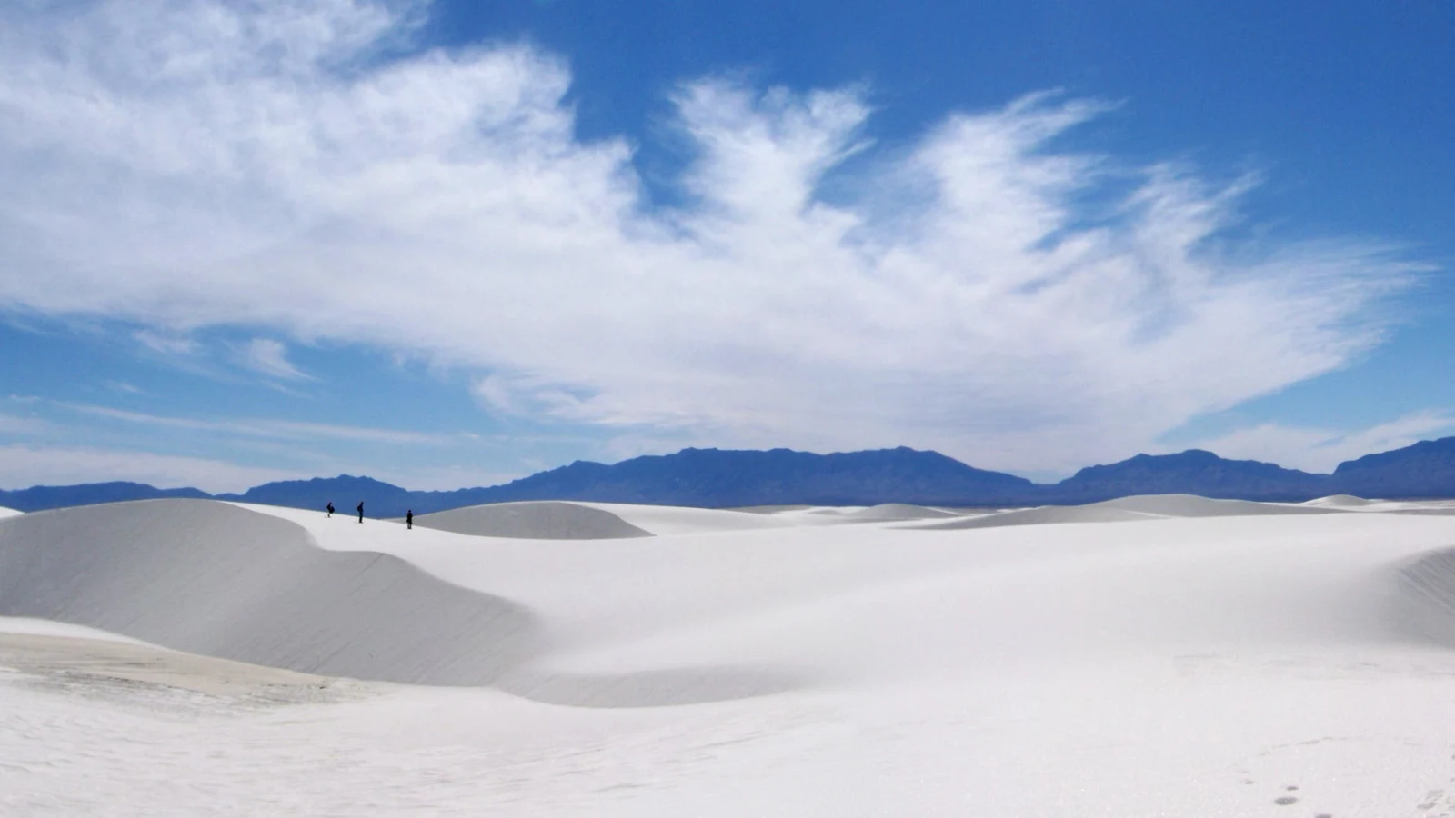 Hikers crossing bright white gypsum dunes on the Alkali Flat Trail, with mountains on the horizon