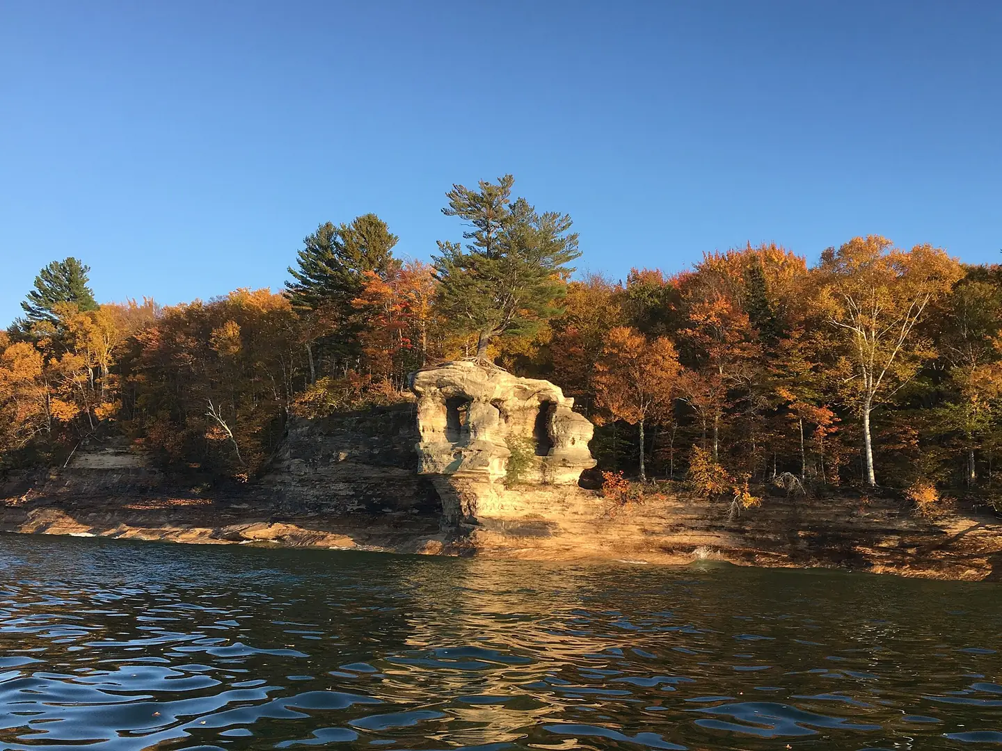 Chapel Rock, a sandstone tower with a lone white pine on top, seen from Lake Superior with fall-colored trees on the shore