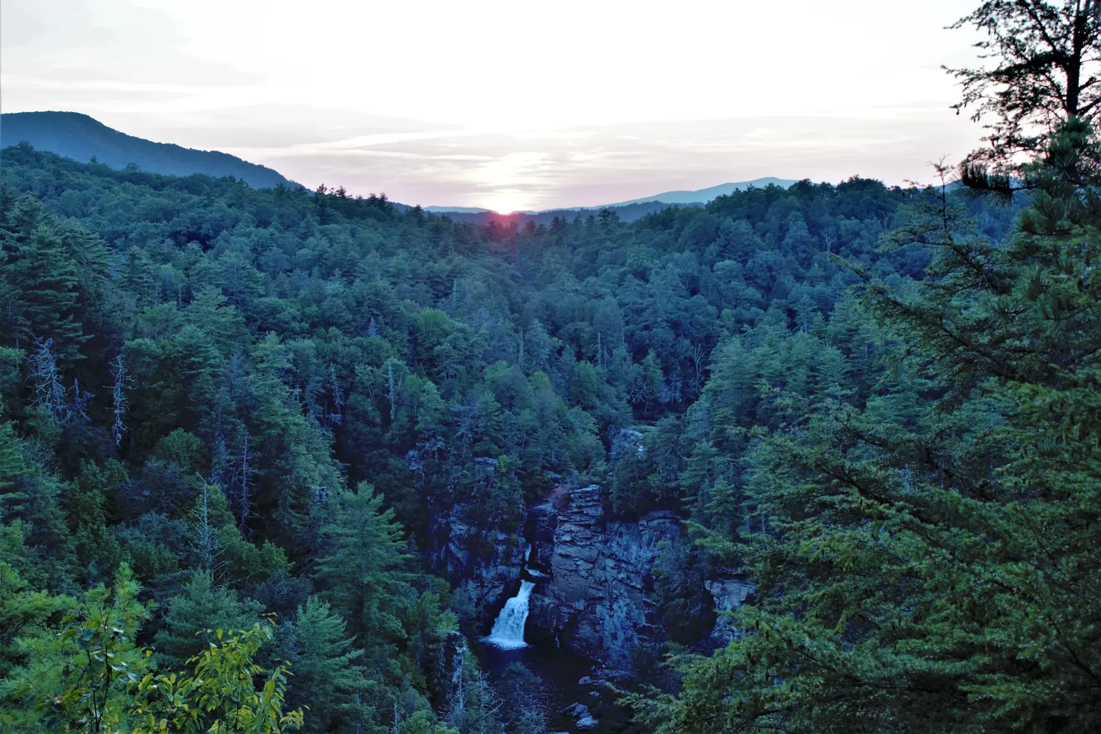 Distant view of Linville Falls in a forested gorge at sunset, with mountain ridges on the horizon